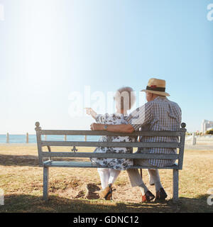 Vue arrière du senior couple relaxing sur un banc avec femme pointant vers la mer. A pris sa retraite l'homme et la femme assise sur un banc à l'extérieur Banque D'Images