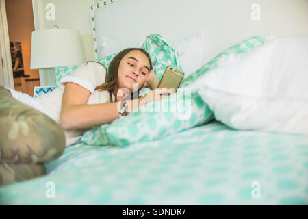 Teenage girl lying on bed reading textes smartphone Banque D'Images