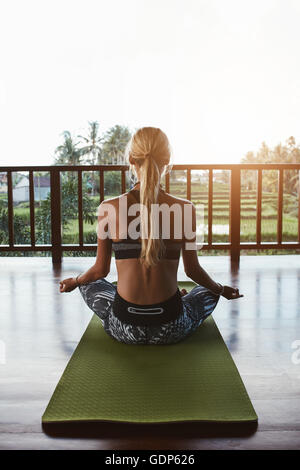 Vue arrière shot of young woman sitting in lotus pose sur tapis d'exercice. Ce modèle féminin de remise en forme à la méditation d'un club de santé. Banque D'Images