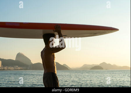 RIO DE JANEIRO - 5 avril, 2016 : un jeune homme brésilien sur la plage de Copacabana, équilibre un surf sur sa tête au lever du soleil. Banque D'Images