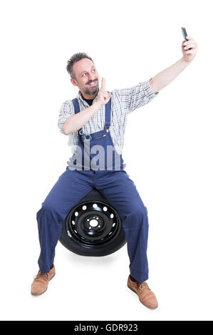 Mécanicien joyeux assis sur le pneu et la prendre avec le smartphone et selfies faisant signe de victoire isolé sur fond blanc Banque D'Images