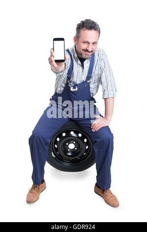 Beau mécanicien confiant assis sur la roue de voiture et montrant cellphone avec écran blanc avec copie espace salon isolated on white Banque D'Images