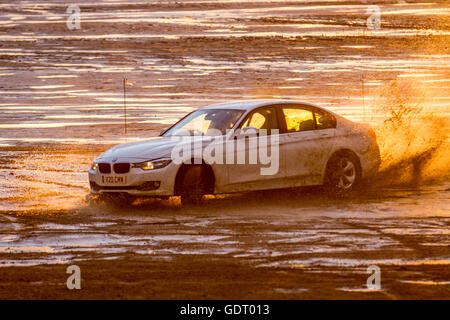 2012 BMW 1598cc berline essence conduite sur la plage, après une forte averse de pluie, à la tombée de la nuit alors que les pilotes de voiture locaux affûtent leurs compétences en pratiquant des virages à main sur le sable humide comme le soleil couchant est reflété dans les piscines de plage à basse eau. Des groupes de « Boy Racers » ont fait monter et descendre les estran lumineux avec leurs voitures bien équipées et très performantes, en effectuant des beignes et d'autres manœuvres dangereuses à grande vitesse. Banque D'Images