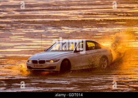 2012 BMW 1598cc berline essence conduite sur la plage, après une forte averse de pluie, à la tombée de la nuit alors que les pilotes de voiture locaux affûtent leurs compétences en pratiquant des virages à main sur le sable humide comme le soleil couchant est reflété dans les piscines de plage à basse eau. Des groupes de « Boy Racers » ont fait monter et descendre les estran lumineux avec leurs voitures bien équipées et très performantes, en effectuant des beignes et d'autres manœuvres dangereuses à grande vitesse. Banque D'Images