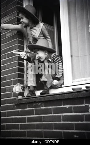 Années 1960, historiques, deux jeunes enfants vêtus de salopettes et de chapeaux de se pencher à l'ouverture d'une fenêtre appartement prétendant être des cowboys, Angleterre. Banque D'Images