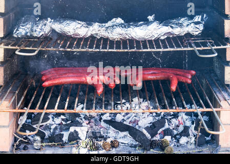 Près d'un barbecue avec des saucisses et pommes de terre au four enveloppé d'aluminium sur un grill Banque D'Images