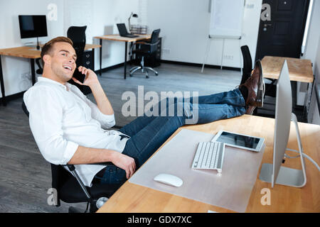 Happy young businessman sitting détendue avec les jambes sur la table et talking on cell phone in office Banque D'Images