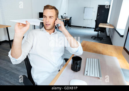 Heureux jeune homme espiègle talking on mobile phone and throwing paper plane in office Banque D'Images