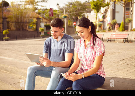 Jeune couple assis sur une place de la ville sur une pierre l'étape à la tablette et à leur téléphone cellulaire avec des sourires à pleines dents heureux Banque D'Images