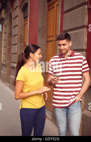 Jeune couple looking at cell phone, bien qu'il partage une blague et souriant joyeusement, habillé par hasard à t-shirts avec des vieux bâtiments beh Banque D'Images