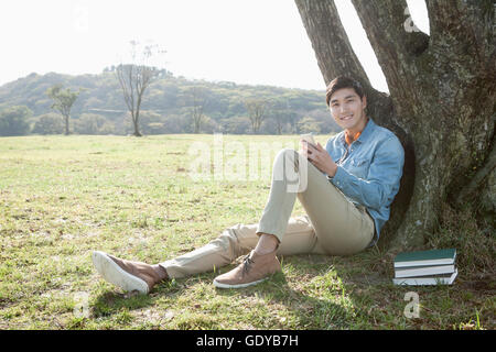 Young smiling man sitting et de repos sous un arbre sur les herbages à regarder/ Banque D'Images