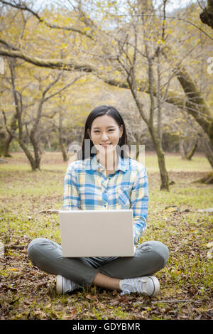 Jeune femme souriante avec un ordinateur portable assis dans un parc à regarder/ Banque D'Images