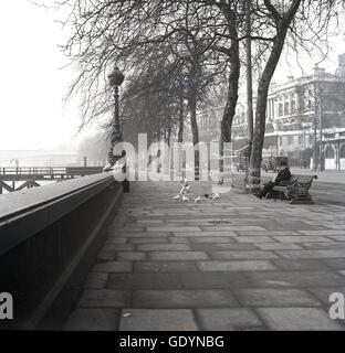 Années 1950, historique, tôt le matin et un homme de ville dans un chapeau de lanceur assis sur un banc de trottoir nourrissant des pigeons sur un remblai de Victoria déserté à côté de la Tamise, Westminster, Londres, Angleterre, Royaume-Uni. Banque D'Images