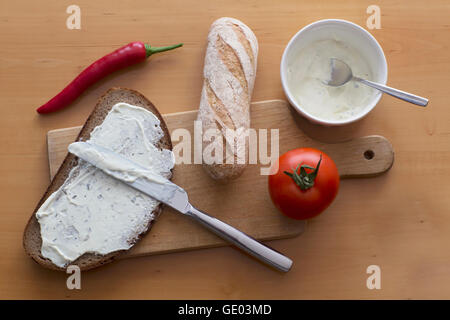 Répandre le fromage sur la tranche de pain gris avec baguette et tomate sur planche à découper Banque D'Images