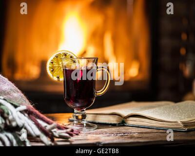 Vin chaud et un livre sur la table en bois. Cheminée avec feu chaud sur l'arrière-plan. Banque D'Images
