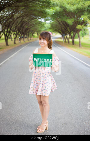 Cute woman hand holding green board panneau avec texte ' ' Bienvenue sur route et arbre, modèle femme. Banque D'Images