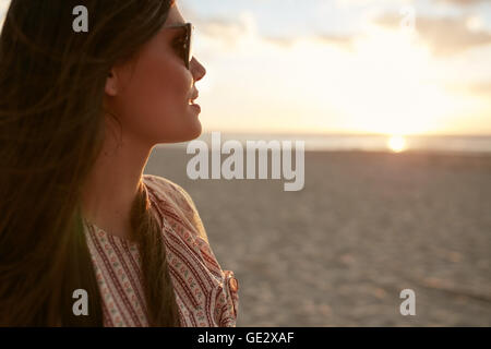 Gros plan de jeunes femmes avec des lunettes de soleil à la recherche au coucher du soleil sur la plage. Modèle féminin sur le bord de la mer au coucher du soleil Banque D'Images