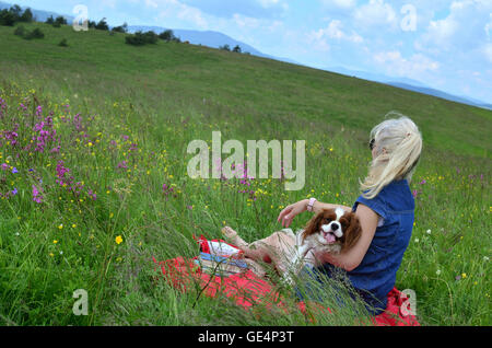 Femme blonde avec son chien détente sur un pré entouré de fleurs roses et l'observation d'un paysage vallonné Banque D'Images