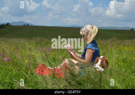 Femme avec chien détente sur un pré entouré de fleurs roses et d'écrire quelque chose dans son calepin, sur jour de vent Banque D'Images
