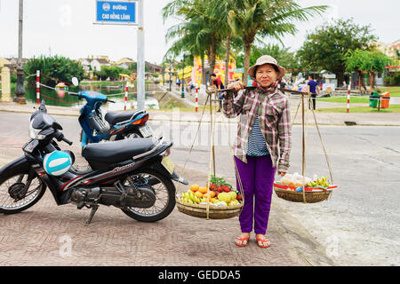 Hoi An, Vietnam - 16 Février 2016 : Asian woman jardiniers portant des fruits dans des bols sur ses épaules dans la rue à Hoi An, au Vietnam. Ramboutans, mangue, banane et mandarin. Banque D'Images