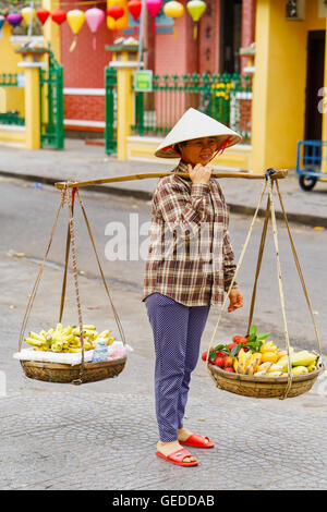 Hoi An, Vietnam - 17 Février 2016 : vendeur transportant des fruits dans des bols sur ses épaules dans la rue à Hoi An, au Vietnam. Ramboutan, mangue et banane. Banque D'Images