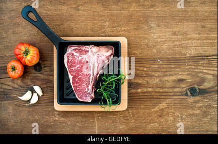 La viande non cuite brutes t-bone steak à l'ail, les tomates, le romarin dans petite casserole foncé sur fond de bois rustique, vue du dessus, copy space Banque D'Images