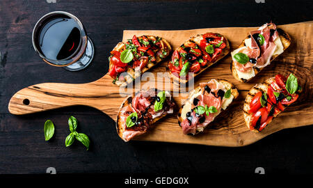 Jeu de Brushetta et verre de vin rouge. Petits sandwiches au jambon, tomates, fromage parmesan, basilic frais, crème balsamique sur planche de bois rustique Banque D'Images