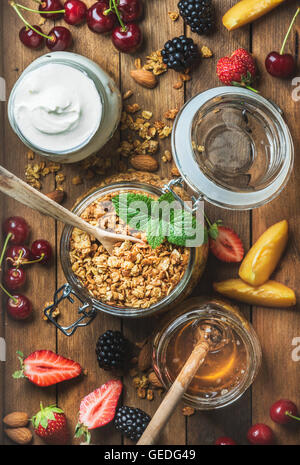 Petit-déjeuner sain ingrédients. Granola avoine en pot ouvert, yaourt et miel servi avec des baies, des noix, des feuilles de menthe fraîche sur fond de bois rustique. Vue de dessus, la composition verticale Banque D'Images