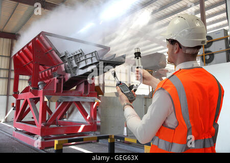 Un ingénieur de l'environnement surveille les niveaux de poussière à côté d'une trémie de matériaux à une usine de récupération de l'aluminium. Photomontage. Banque D'Images