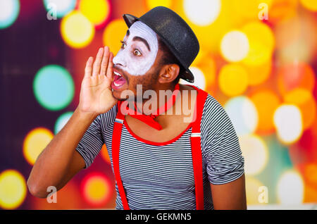 Headshot homme pantomime avec peinture faciale posant pour la caméra à l'aide de mains autour de la bouche en criant, floue lumières fond Banque D'Images