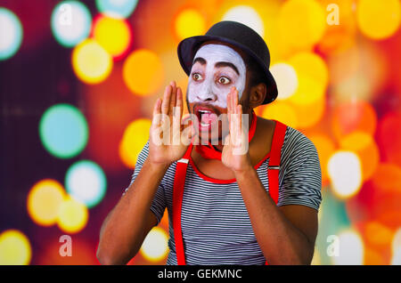 Headshot homme pantomime avec peinture faciale posant pour la caméra à l'aide de mains autour de la bouche en criant, floue lumières fond Banque D'Images