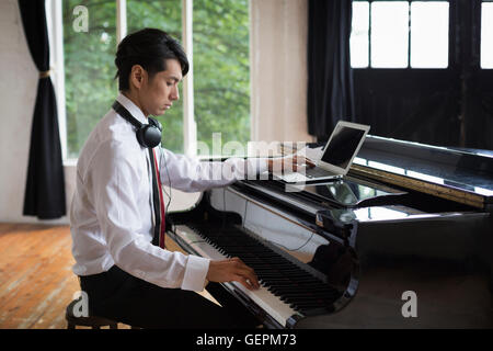 Jeune homme assis à un piano dans un studio de répétition, jouant et simultanément à l'aide d'un ordinateur portable. Banque D'Images