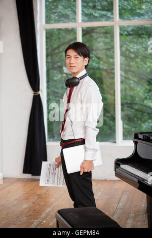 Jeune homme debout à côté d'un grand piano dans un studio de répétition, tenant des partitions de musique. Banque D'Images