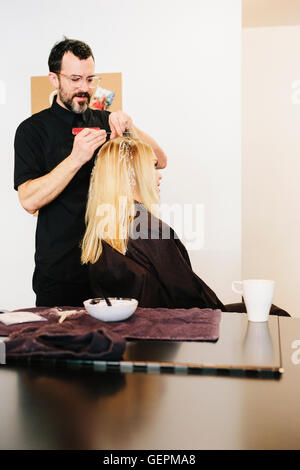 Un coloristees travailler avec foils de donner à un client avec de longs cheveux blonds les temps forts et l'utilisation des couleurs. Banque D'Images