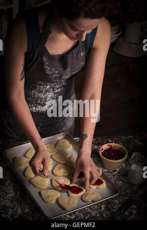 La Saint-Valentin, jeune femme debout dans une cuisine, la diffusion de la confiture de framboises sur les biscuits en forme de coeur. Banque D'Images