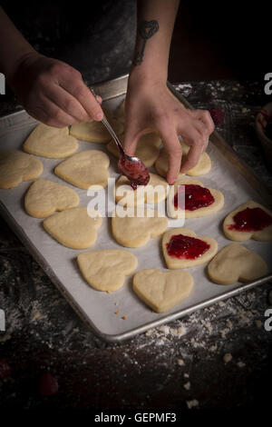 La Saint-Valentin, femme répandre la confiture de framboises sur les biscuits en forme de coeur. Banque D'Images