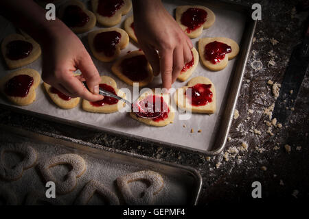 La Saint-Valentin, femme répandre la confiture de framboises sur les biscuits en forme de coeur. Banque D'Images