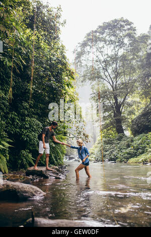 Piscine shot of young man helping woman crossing stream. En traversant la forêt couple creek. Banque D'Images