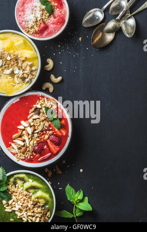 Petit-déjeuner d'été en bonne santé concept. Smoothie aux fruits colorés avec des écrous, des bols de céréales d'avoine et de feuilles de menthe, 5 cuillères d'argent sur fond noir. Jaune, rose, rouge, vert. Vue de dessus, copy space Banque D'Images