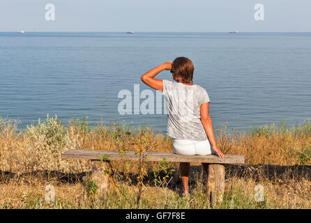 Femme d'âge moyen blanc à poils courts en t-shirt, short et lunettes de soleil assis sur le vieux banc en bois avec Black Sea seascape Banque D'Images