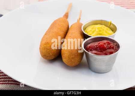 Deux chiens de maïs avec la sauce métal tasses contenant de la moutarde et du ketchup sur une plaque blanche. Banque D'Images