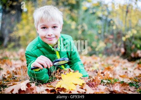 Parution du modèle. Boy holding loupe sur feuille d'automne, smiling, portrait. Banque D'Images