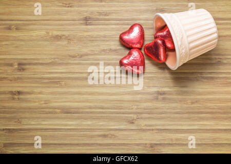 Des bonbons en forme de cœur enveloppé dans un papier rouge brillant couché dans vase en céramique sur la texture en bois. Arrière-plan de thèmes romantiques. Banque D'Images