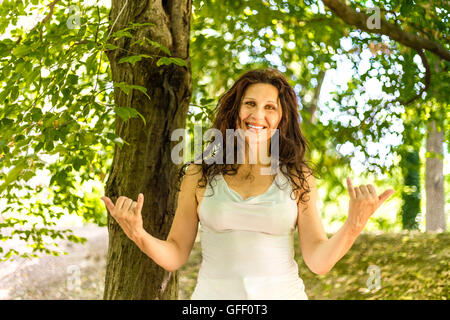 Close up of young woman smiling classy busty à l'appareil photo tout en faisant bienvenue signe shaka contre green garden background with copy space Banque D'Images