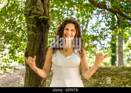 Close up of young woman smiling classy busty à l'appareil photo tout en faisant bienvenue signe shaka contre green garden background with copy space Banque D'Images