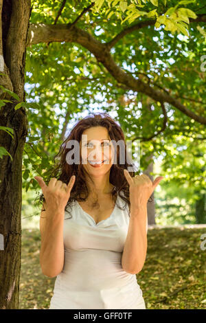 Close up of young woman smiling classy busty à l'appareil photo tout en faisant bienvenue signe shaka contre green garden background with copy space Banque D'Images