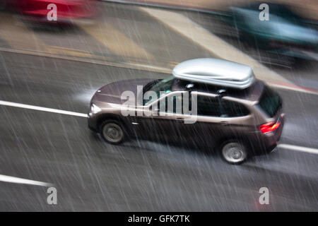 Jour de pluie dans la ville : une voiture, conduite avec une boîte de rangement sur le toit, dans la rue, frappé par la forte pluie de grêle, en mouvement b Banque D'Images