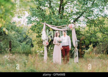 Tenue de mariage Couple avec bouquet de fleurs et de verdure dans jardin, arch on background Banque D'Images