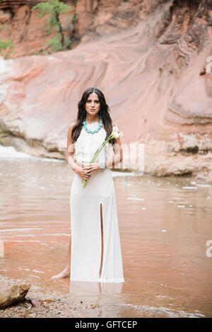 Jeune femme aux longs cheveux bruns portant une longue robe blanche debout par une rivière holding Arums Banque D'Images