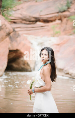 Jeune femme aux longs cheveux bruns portant une longue robe blanche debout par une rivière holding Arums Banque D'Images